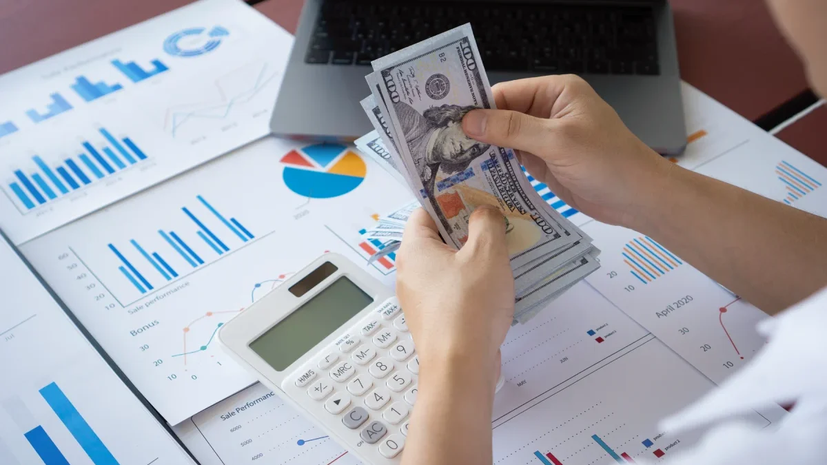 Business owner counting money over financial reports and charts representing cash flow management and business profitability.