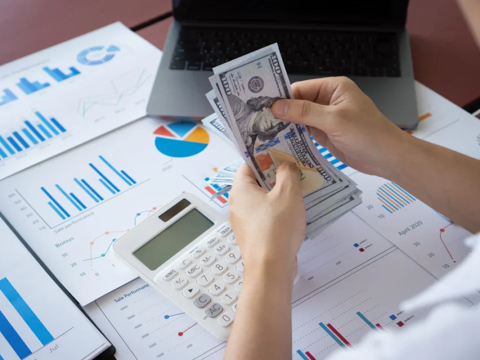 Business owner counting money over financial reports and charts representing cash flow management and business profitability.